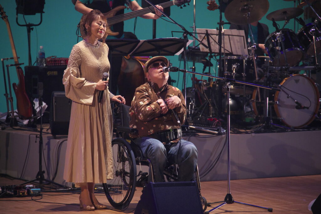車椅子で全盲のハーモニスト山下純一さんのよこで一緒に歌う麻生さん。